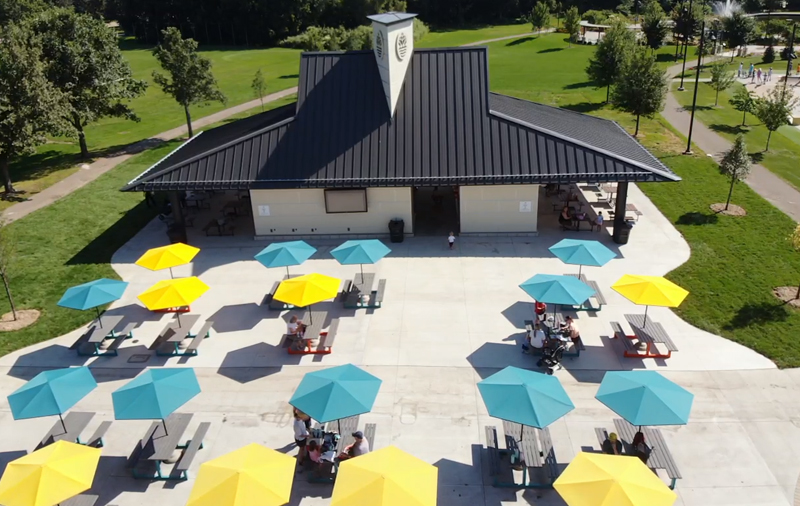 Aerial view of the Shoreview Commons park pavilion in Shoreview, Minnesota, featuring a covered seating area with colorful patio umbrellas, tables, and surrounding green space.