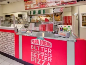 Interior of a Papa John's franchise restaurant featuring a service counter, pizza preparation area, and branded signage.
